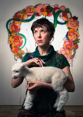 Artist Peregrine Honig with a taxidermied lamb in front of one of her recent paintings in the Unicorn exhibit.